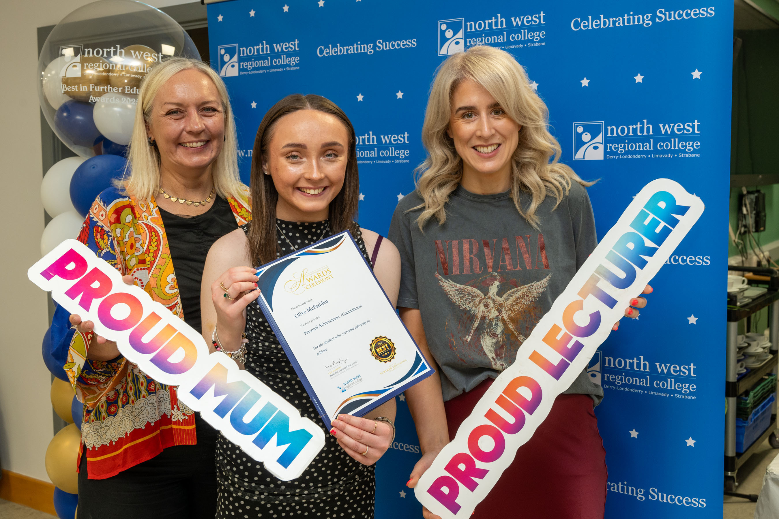 Two people at celebration event. One is holding a sign that says proud mum, and another proud lecturer