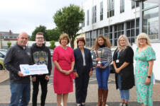 Seven people standing outside including young black female holding an award and an older male holding the college logo prop