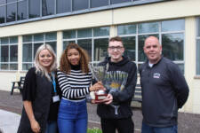 Two students outside college building holding a large trophy with two staff members