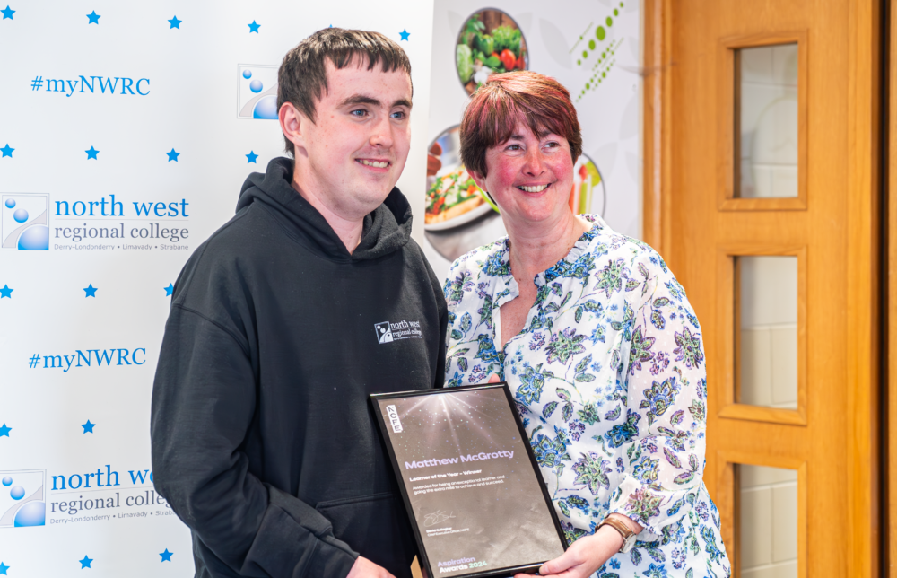 A male student and female lecturer smile looking away from the camera. They are holding a framed award.