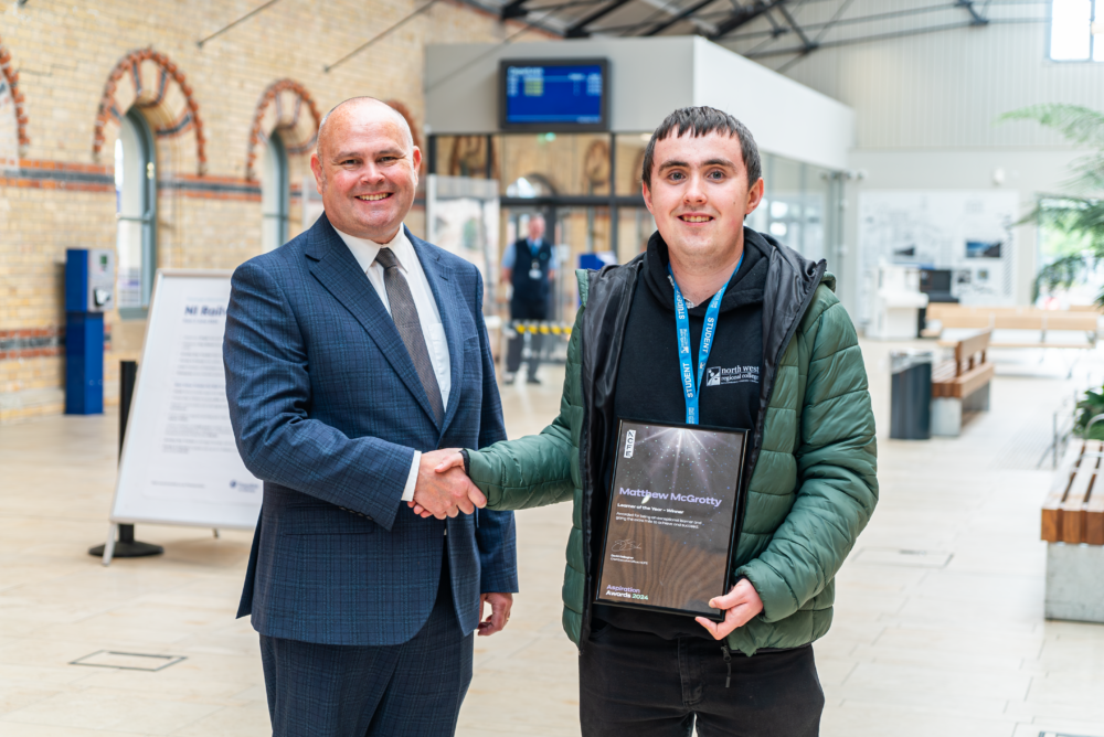 A man in a suit, and a student in a hoodie on the platform of a railway station with a train behind them.