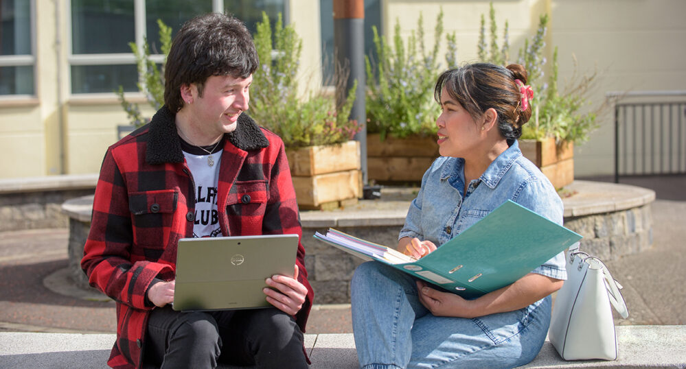 Male in red checked jacket holding laptop smiles at female Filipino holding a blue folder