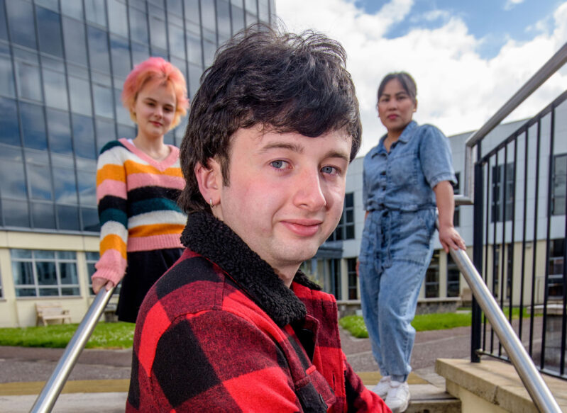 Young man wearing a red check jacket with a young female with pink hair and Filipino woman in background standing on steps