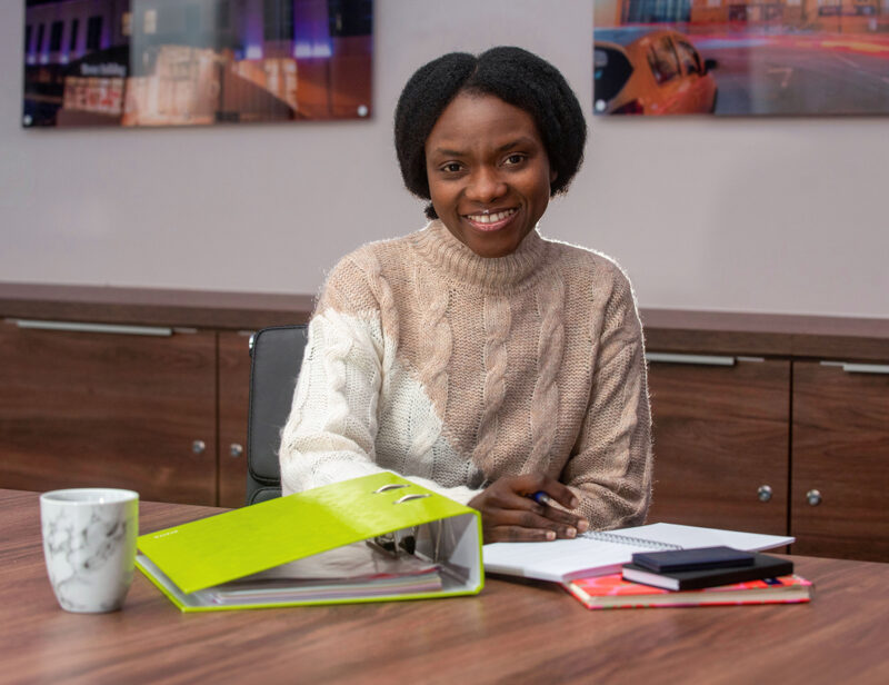 Black woman in cream jumper sitting at desk with cup, books and green lever arch folder in front of her and photographs on the wall behind her