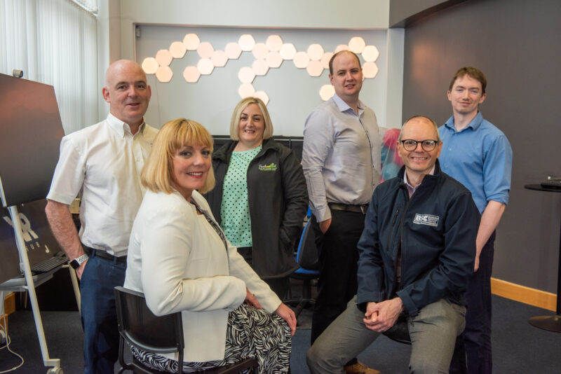 Six people smile at the camera. They are pictured in a room with bright hexagons on the wall.