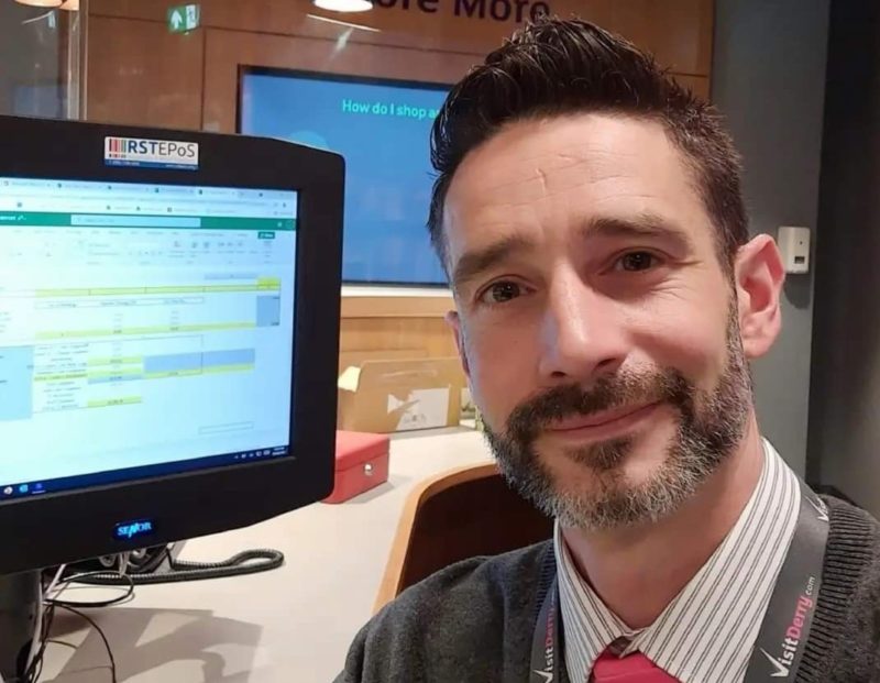 Man with a beard smiling. He is sitting in an office in front of a computer