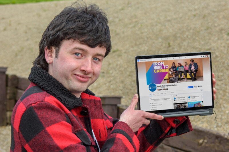 Male student holding a laptop. He is wearing a checked coat and smiling.