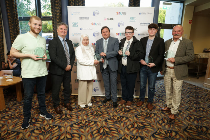 Six men and one women pictured with their prizes at an awards ceremony. There is branding behind the group.
