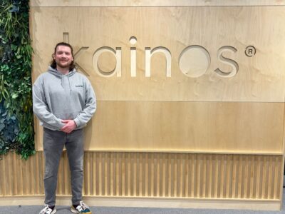Man dressed in grey hoodie with long hair in front of a wooden sign spelling out Kainos.