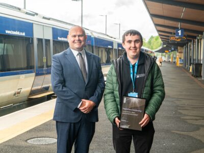 A man in a suit, and a student in a hoodie on the platform of a railway station with a train behind them.