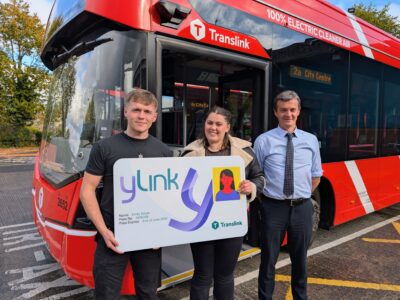 Two students and a bus driver outside standing outside with a bus behind them. They are hold a card with ylink on it