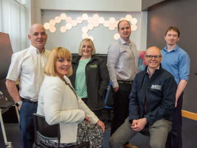 Six people smile at the camera. They are pictured in a room with bright hexagons on the wall.