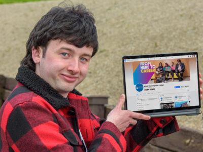 Male student holding a laptop. He is wearing a checked coat and smiling.