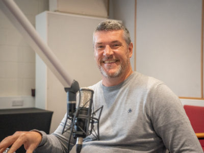Man in grey jumper sitting in front of a radio mic.