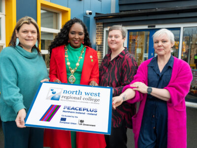 Four women outside a community centre hold a graphic with the NWR and Peaceplus logo