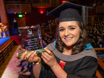 Woman holding a trophy wearing a graduation gown and cape. She is standing in front of a crowd in an auditorium.