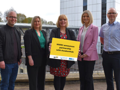 Two men and three women standing outside a college building. One of the women is holding a sign which states NWRC welcomes partnership with Awakenhub