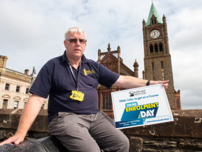 Man sitting on Derry's walls holding a sign about enrolment day. The guildhall is in the background,