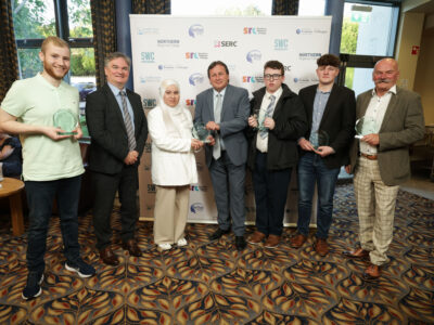 Six men and one women pictured with their prizes at an awards ceremony. There is branding behind the group.