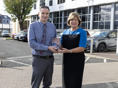 Man and woman outside a college building. They are smiling and holding a crystal award.