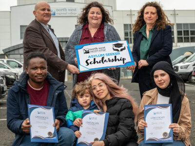 seven people standing in two rows outside a college building with cars parked at both sides. The back row of three people are holding a sign 'NWRC Asylum Seeker Scholarship' and the front row are holding certificates.