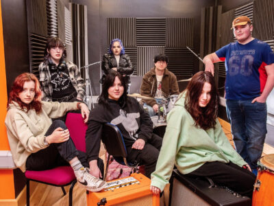 Seven students in a music studio surrounded by drums, speakers and microphones