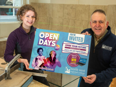 Rebecca and David hold a prop with Open Day on it. They are standing in a tiling workshop