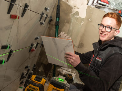 Student with glasses pictured in a house where he is tiling a bathroom