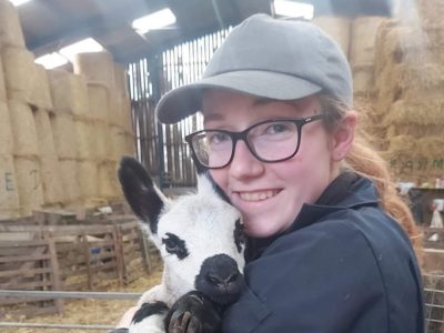 Woman in a baseball cap standing in a barn holding a baby calf. She is wearing glasses and smiling